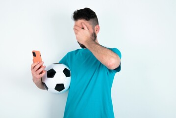 Young man holding a ball over white background looking at smart phone feeling sad holding hand on face.