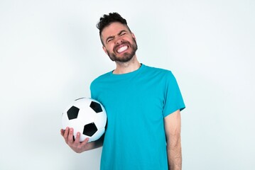Positive Young man holding a ball over white background with overjoyed expression closes eyes and laughs shows white perfect teeth