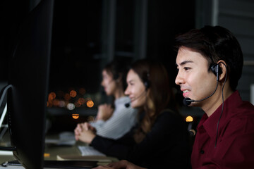 Happy smiling Asian man work at call center service consultant with teammates at night, night shift customer service executive staff with microphone headset use computer for support customer at night.