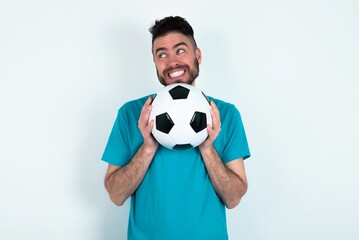 Happy Young man holding a ball over white background anticipates something awesome happen, looks happily aside, keeps hands together near face, has glad expression.