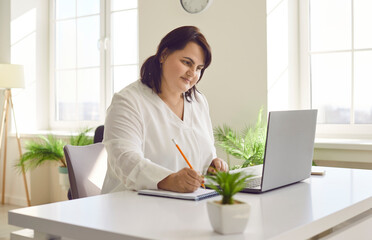 Overweight brunette young woman using laptop in modern office. Focused plus size woman reviewing...
