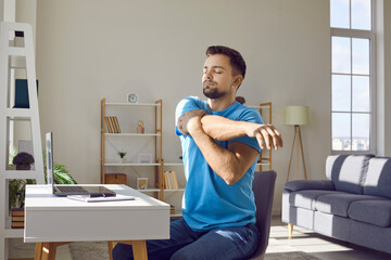 Man stretches during work break. Young man in blue T shirt sitting by desk with laptop computer in...