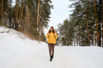 Young woman traveler with  backpack and yellow coat walking on snow covered road in winter forest in frosty weather.