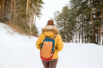 Young woman traveler with  backpack and yellow coat walking on snow covered road in winter forest in frosty weather.