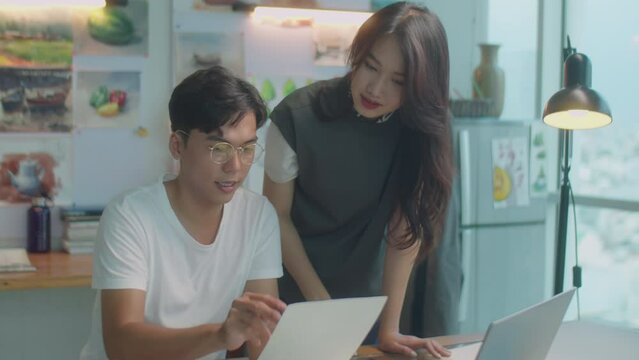 Medium shot of young Asian colleagues discussing paper drawing and using laptop while working on creative project in art studio