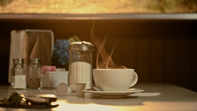 A steaming hot cup and saucer of coffee with steam rising as the light from the sun rises through the dirty window of a diner.