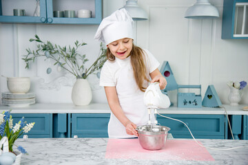 little cute funny smiling girl wearing a white chef's hat and uniform  shaking egg whites with mixer in the blue and white kitchen with easter decoration