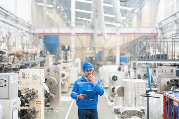 European white male engineer Powering the radio to do business and walking in the industrial factory Around there are machines working. He wore a uniform and helmet.