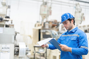 European white male engineer in the industrial plant He was carefully checking the list of items from the list of items. There were machines working around him. Wear uniforms and wear helmets.