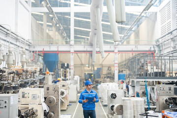 European white male engineer Was walking to explore the system in the plastic industry factory. There are radio communications and list notes. around there are machines He wore a uniform and helmet.