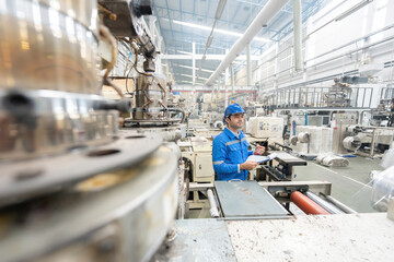 European white male engineer standing working in the factory Holding a walkie-talkie and a listnote. Around there are machines working. Wear uniforms and wear helmets.