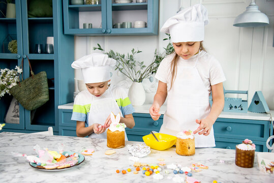 two little cute funny smiling kids wearing a white chef's hats and uniform decorating easter bread with food coloring  in the blue and white kitchen with easter decorations