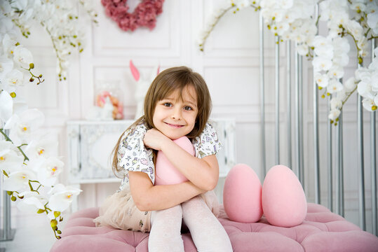 little funny cute smiling brunette girl sitting on oink sofa and playing with three big pink toy easter egg in room with white orchids