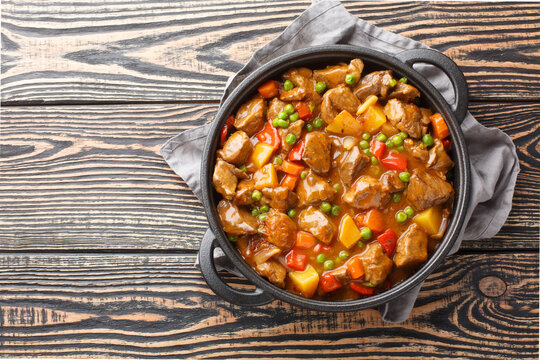 Spicy Beef Cooked With Potatoes, Carrots, Tomatoes, Green Peas, Onions In Their Own Juice Close-up In A Saucepan On A Wooden Table. Horizontal Top View From Above