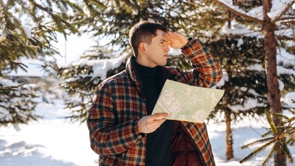 A man standing in the middle of a winter forest looks into the distance, shielding his eyes from the sun. A brunette man with a map in his hands stands in the middle of a snowy forest on a sunny day.