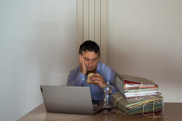 Image of a desperate man holding a sandwich and a small bottle of water who has to work overtime in his office due to overwork.