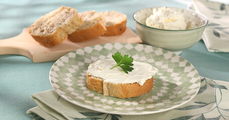 slice of bread with cream cheese. bowl with creamy spread and sliced bread on a green tablecloth. healthy breakfast on a sunny day.