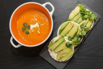 Pumpkin and carrot cream soup, round crispy rice crackers with avocado on black board background.