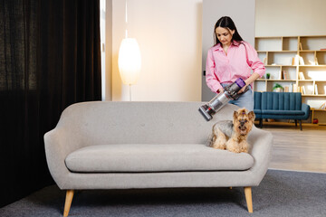 A cute young woman is vacuuming a couch with her cute dog sitting on it with a cordless vacuum...