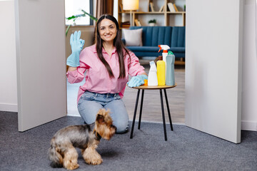 Young woman sitting near the table with cleaning products at home and her little dog is running next to her