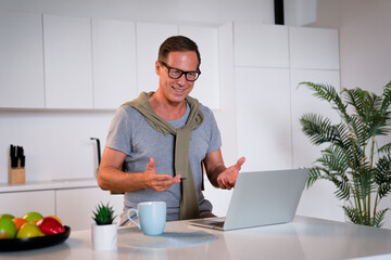 mature man using laptop, making video call, sitting at table in kitchen, senior teacher mentor...
