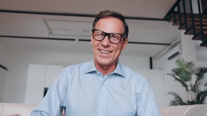 Happy mature 60s man in glasses waving hello at camera, smiling, laughing, speaking on video call at home