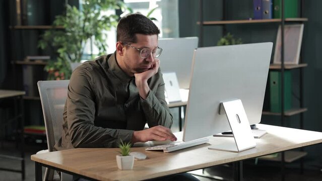 young caucasian businessman in casual shirt and glasses using computer at workplace. looking bored using pc, leaning head on hand