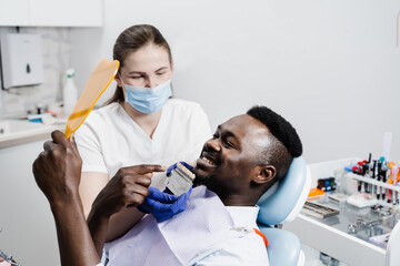 Dentistry. Dentist showing teeth color shades guide for tooth whitening for african patient in dental clinic. African is looking at veneers or implants teeth color matching samples in doctor hands.