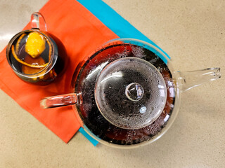Glass transparent teapot with hot tea on colored napkin on a table top. Drops of condensate on inner surface. Cup of tea with lemon as a drink flavoring in background. View from above. Selective focus