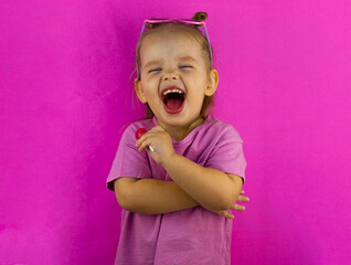 Cute baby girl laughs at the top of her mouth with a lollipop in her hand on a pink background. A...