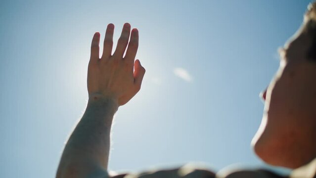 Man closing sun by hand, blue sky, covering sunlight with arm. Young person looking at bright light.