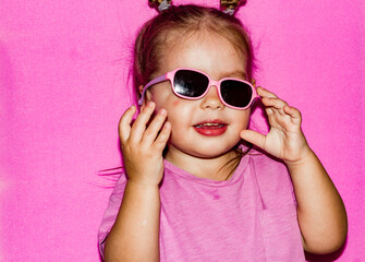 Humor. On a pink background, a little girl child funny adjusts her sunglasses that have fallen off. The child is funny trying to return the sunglasses that have fallen into place.