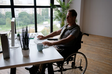 An elderly man in a wheelchair works in the office at the computer. Silhouette of a man