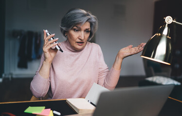 Unsure mature woman sitting with laptop and smartphone