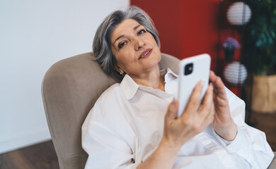 Smiling mature woman resting with smartphone on sofa
