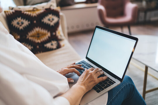 Crop aged woman typing on laptop while sitting on sofa