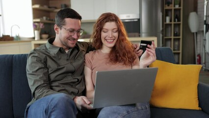young happy loving couple in home indoors on sofa using laptop computer holding credit card.