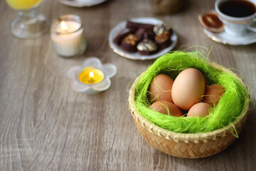 Easter eggs in the basket, bowl of cookies, chocolate pralines, Easter bunny figurine, cups of tea, glasses of juice, flowers and lit candles on the table. Selective focus.