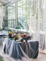 Delicate wedding decor of a banquet in a greenhouse. Panoramic windows, plants hanging from the ceiling, white tulle, glass balls. Bouquets of colorful flowers on the table.