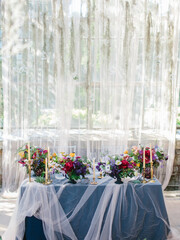 Delicate wedding decor of a banquet in a greenhouse. Panoramic windows, plants hanging from the ceiling, white tulle, glass balls. Bouquets of colorful flowers on the table.