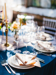 Wedding banquet table setting in blue and white. In the foreground are white plates with napkins, wine glasses, knives and forks. Behind bright flowers. Drapery with tulle.