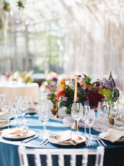 Wedding banquet table setting in blue and white. In the foreground are white plates with napkins, wine glasses, knives and forks. Behind bright flowers. Drapery with tulle.