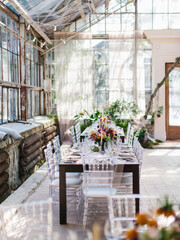 Serving a wedding banquet in the greenhouse. In the center of the table are bouquets of flowers, candles in golden candlesticks, dishes. transparent chairs. Panoramic windows, green plants.