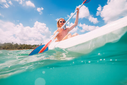 Underwater view angle to a smiling blonde teenage boy in fashion sunglasses rowing single sit kayak with palm grove background. Active people summer vacation sea concept image, Le Morne, Mauritius.