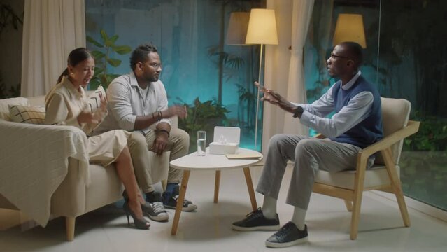 Full shot of diverse family couple sitting on couch and arguing while African American psychologist trying to resolve their conflict during therapy session