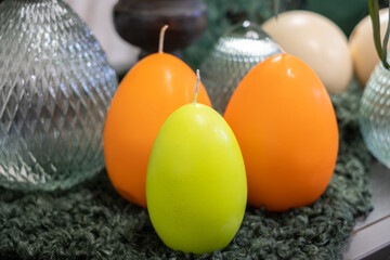 Fototapeta na wymiar Beautiful, bright, orange and yellow candles like Easter eggs on a fluffy dark green napkin. Glass transparent decorative Easter eggs in the background