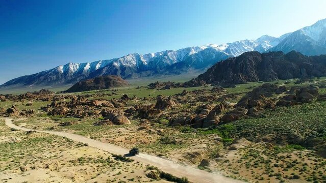 Aerial Panning Shot Of Adventure Vehicle Moving In Desert Area By Snowcapped Mountain Range, Drone Flying Forward Over Plants On Sunny Day - Alabama Hills, California