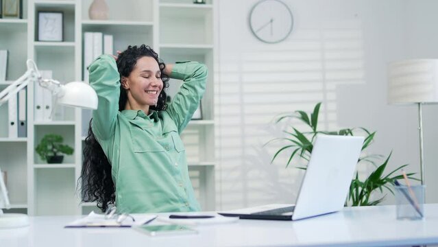 Happy young hispanic employee typing with laptop then relaxing stretching arms smiling in office during enjoyable work break People workplace rest relaxation after successfull complet work