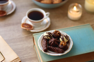Books, reading glasses, e-reader, plate of chocolate pralines, bowl of cookies, cups of tea and lit candles on the table. Selective focus.
