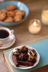Books, reading glasses, e-reader, plate of chocolate pralines, bowl of cookies, cups of tea and lit candles on the table. Selective focus.